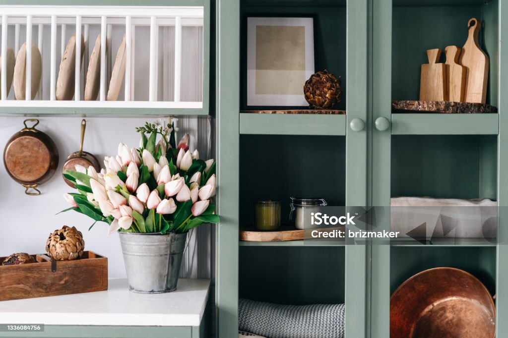 Green vintage kitchen cupboard with glass doors Green vintage cupboard with glass doors, set of various sized wooden cutting boards and tablecloths, countertop with white surface, lots of flowers in metal bucket, comfortable kitchen enviroment Kitchen Stock Photo