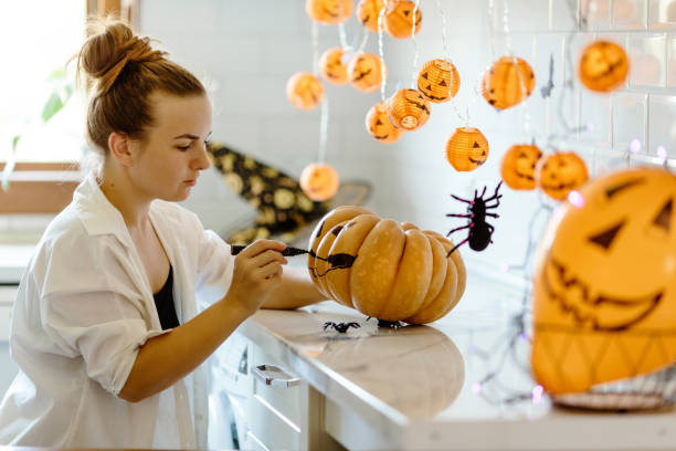 mujer pinta la cara en calabaza naranja para halloween. hojas de otoño. decoraciones de fiestas de otoño. sentado detrás de una mesa en la habitación decorada con temas de halloween, sosteniendo calabazas pintadas a mano - halloween pumpkin party carving fotografías e imágenes de stock