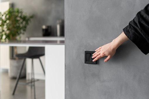 Cropped shot of woman turning light on or off in kitchen using black switch located on grey wall, modern kitchen interior design with plants, furniture and appliances in blurred background