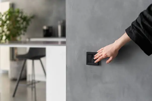Cropped shot of woman turning light on or off in kitchen using black switch located on grey wall, modern kitchen interior design with plants, furniture and appliances in blurred background