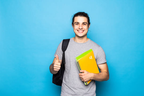 istock Happy student, holding his backpack and books, standing raising his thumb up, standing on blue background. 1336098394