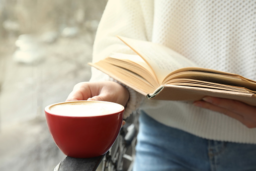 Woman with cup of coffee reading book outdoors, closeup