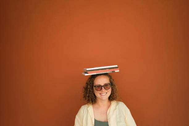 頭の上に本を持つ楽観的な女の子。オレンジ色の背景。虚空。本を持つ眼鏡をかけたカジュアルな服装の若い女性に笑顔。スペースをコピーする - balance book university glasses ストックフォトと画像