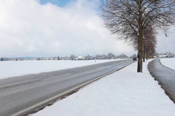 Winterly tree lined country road in the Bavarian Alps. A car drives slowly on the slush covered road.