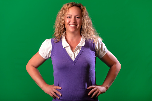 Front portrait of mature blonde woman on green background. She has white blouse with purple vest, happy smiling and looking at camera. She has hands on hips.