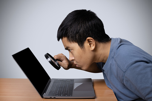 Young man observing laptop with magnifying glass isolated on gray background.
