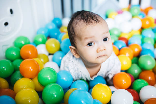 Happy asian laughing baby boy having fun in ball pit on birthday party in kids amusement park and indoor play center and looking at camera. Photo of Child playing with colorful balls in playground ball pool during the day. Activity toys for little kid.