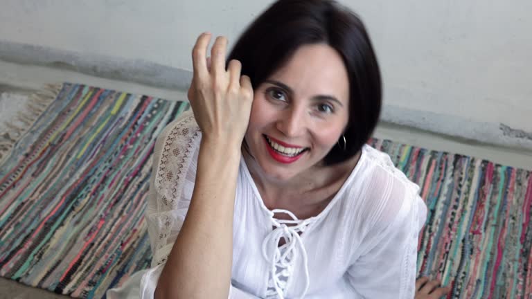 Smiling confident young ethnic woman looking at camera posing alone at home on the ground, happy girl in white dress with red lipstick and facial wrinkles sitting on colorful carpet
