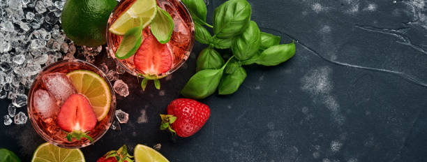 two glasses of punch and fresh ingredients for making lemonade, infused detox water or cocktail. strawberries, lime, mint, basil, ice cubes and shaker on black stone or concrete background. top view. - ice cube cold mint punch imagens e fotografias de stock