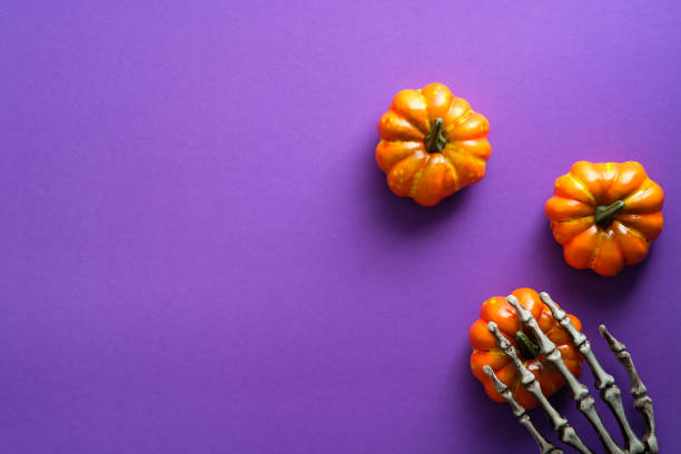 istock Happy Halloween holiday composition with pumpkins and skeleton hand on violet background. Flat lay, top view, copy space. 1336140033