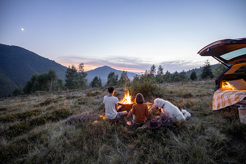 Couple sitting together with a dog by the fire, having romantic picnic, enjoying mountain landscapes on the evening at dusk. Couple traveling by car in nature