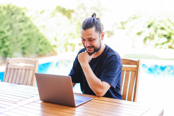 istock Young man using a laptop computer in a garden with a swimming pool. 1336143338