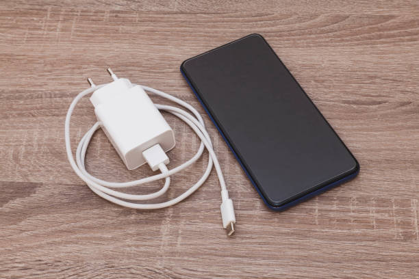 Mobile phone with charger and cable On a wooden table there is a cell phone and a charger with its USB cable. The cable is white and coiled. The smartphone is turned off. battery charger stock pictures, royalty-free photos & images
