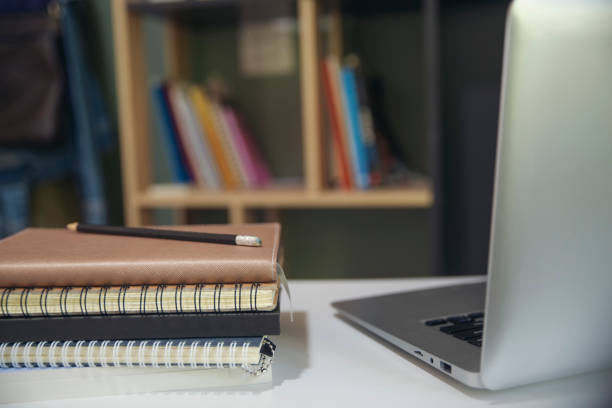 istock Desk for student education concept. Planner Book, laptop, and pencil for study for exam placed on white School table with bookshelf background for student plan to work online at home. 1336149983