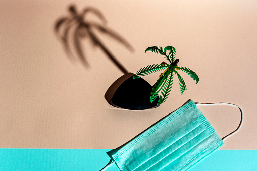palm tree figurine and medical protective mask on a pink and turquoise background, minimalism, vacation at the sea during the pandemic, vacation during covid 19