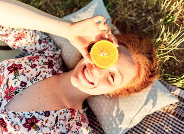 Gorgeous young woman gracefully holds a half orange on her face. She has red hair and is wearing a flowery dress. She is stretched out on a picnic tablecloth in the countryside.