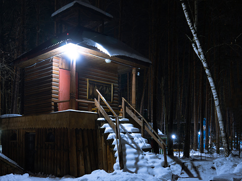Small log house in a winter snow-covered forest at night. The staircase leading to the house is covered with snow. The lantern hangs over the door of the house. Small fortress in the winter forest.