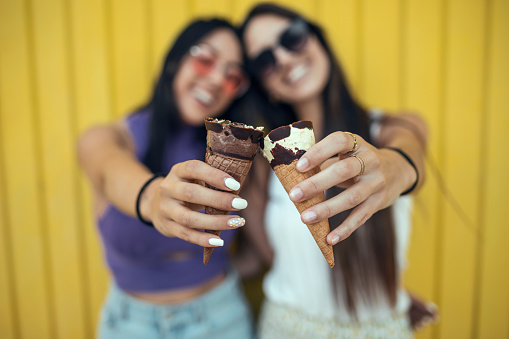 Shoot of two beautiful young women eating ice cream while having fun on yellow background.