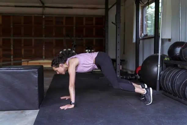 A gorgeous thirty something ethnic woman is exercising inside her home gym. She's wearing a purple tank top and doing burpee box jumps.