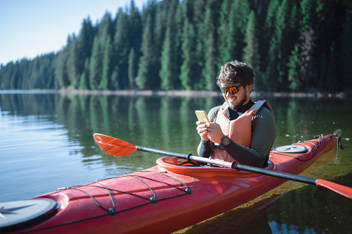 Handsome happy young man using smartphone on kayak in the lake
