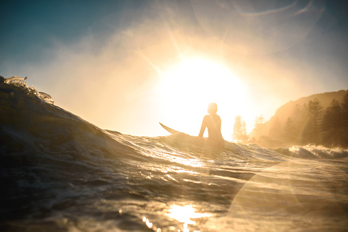 Low angle view with lens flare and specular highlights of silhouetted female surfer standing with her board in shallow water and watching waves at sunrise.