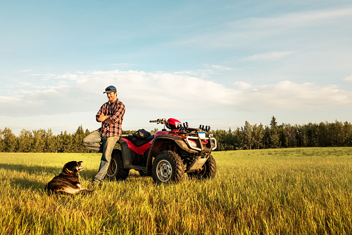 Farmer standing by his quad bike looking at the dog sitting in front on the field on a summer day