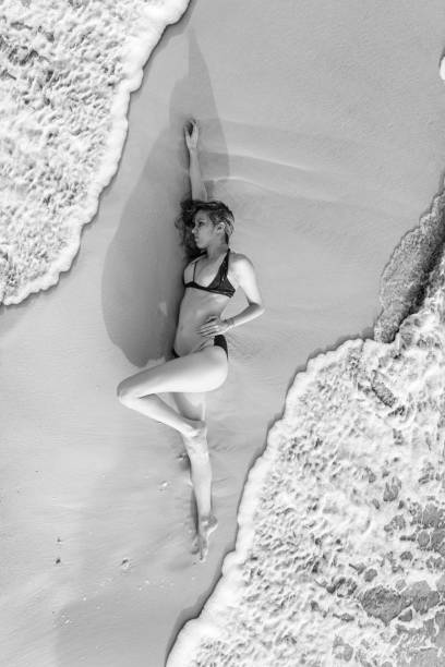 A monochromatic image capturing a woman in a white outfit standing by the water's edge. Her pose conveys a sense of grace and contemplation, against the backdrop of calm waves and an expansive ocean. The composition emphasizes fluidity and elegance, with the subdued colors enhancing the mood of tranquility and introspection.