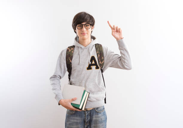 Handsome teen boy students carrying a backpack and books pointing finger standing on white background.