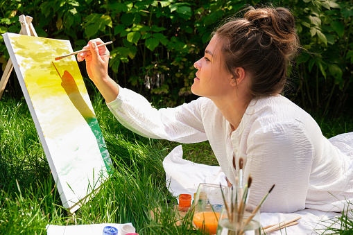 Artist painting on the easel outdoors in the garden. Open air outdoor art workshop. Draw on the canvas with brush and palette sitting on the grass during a picnic in a park. Creativity, art and hobby concept