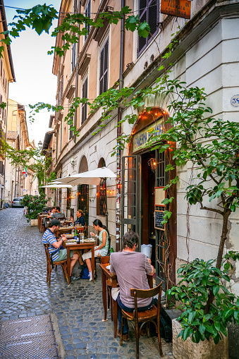 Rome, Italy, July 22 -- Tourists and residents enjoy traditional Roman cuisine sitting in a characteristic and lovely tavern along an alley of the Rione Monti (Monti district), in the historic heart of Rome. The Monti district is a popular and multi-ethnic quarter much loved by the younger generations and tourists for the presence of trendy pubs, fashion shops and restaurants, where you can find the true soul of the Eternal City. This district, located between the Esquiline Hill and the Roman Forum, is also rich in numerous Baroque-style churches and archaeological remains from the Roman era. In 1980 the historic center of Rome was declared a World Heritage Site by Unesco. Image in high definition format.