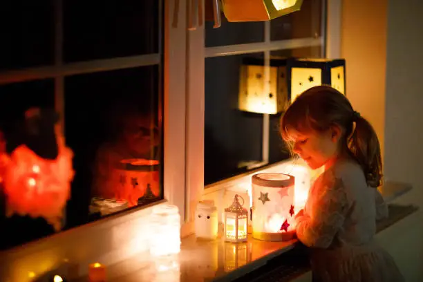 Little kid girl sitting by window with selfmade hand crafted lanterns with candles for St. Martin procession. Toddler child looking at glowing lantern. German tradition Martinsumzug. Home decoration.