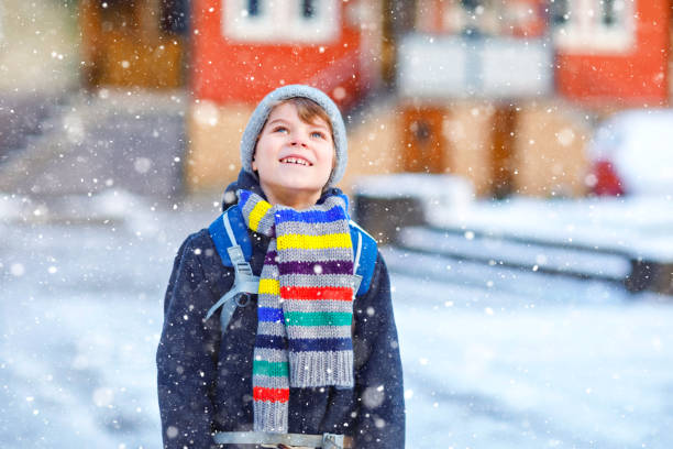 Little school kid boy of elementary class walking to school during snowfall. Happy child having fun and playing with first snow. Student with backpack or satchel in colorful winter clothes