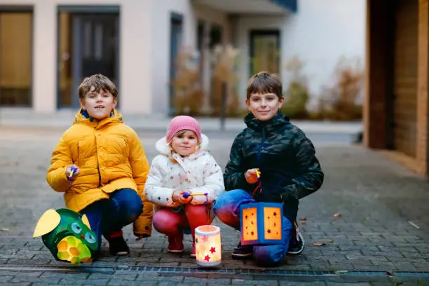 Little toddler girl and two kids boys holding selfmade lanterns with candle for St. Martin procession. Three Healthy children happy about family parade in kindergarten. German tradition Martinsumzug.