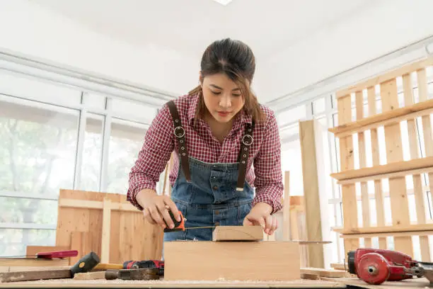 Female carpenter using tape measure while measuring and making notes on woodcraft in the carpentry shop. Handywoman apprentice working in a workshop. DIY woodworking furniture crafts, Hobbies concepts
