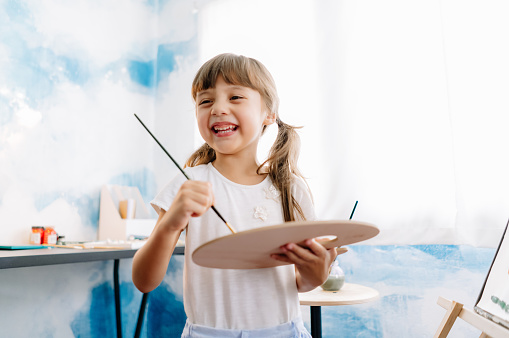 Happy and playful cute blonde girl holding brush and palette in preschool. Children and happiness, Creativity and freedom concept. Arts and crafts.