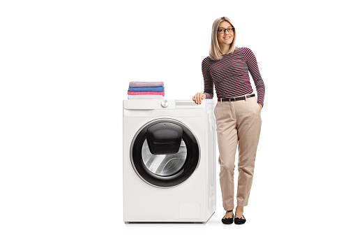Full length portrait of a young woman leaning on a washing machine with folded clothes on top isolated on white background