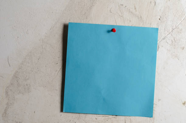 Blue square piece of paper. The note paper is attached with a stationery button. A gray cement wall with scratches. Vertical.