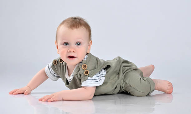 little boy in a green casual overalls, barefoot, lies on the floor and looks with interest into the frame. The kid stopped frozen crawling on the floor