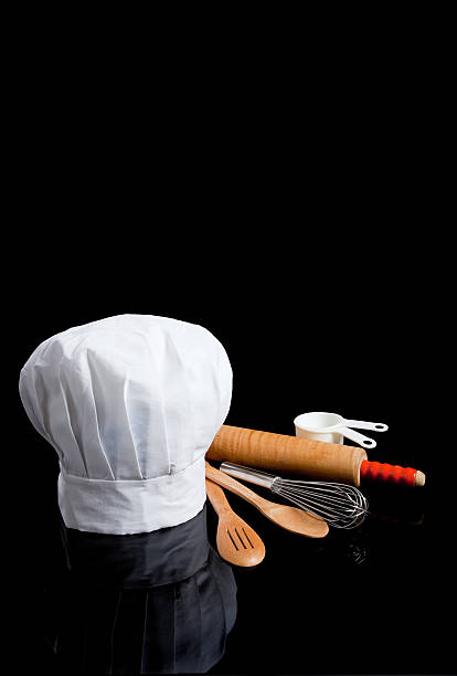 A chef's toque with kitchen utensils including wooden spoons, rolling pin, wire whisk and measuring cups on a black background