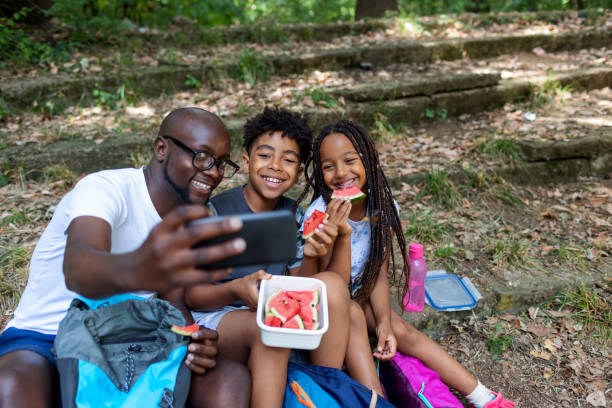 Black Family Making Selfie Father and his two children eating watermelon and making selfie in the woods. black man eating watermelon stock pictures, royalty-free photos & images