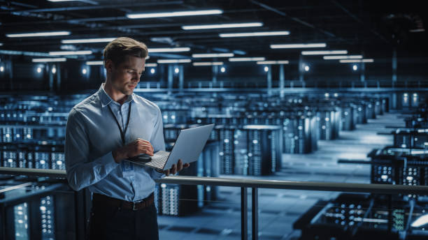 istock Portrait of IT Specialist Uses Laptop in Data Center. Server Farm Cloud Computing Facility with Male Maintenance Administrator Working. Cyber Security and Network Protection. 1336250751