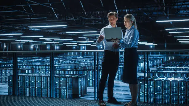 Photo of Data Center Female e-Business Enrepreneur and Male IT Specialist talk, Use Laptop. Two Information Technology Engineers work on Bridge Big Cloud Computing Server Farm.