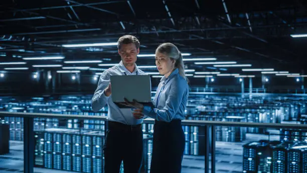 Photo of Data Center Female e-Business Enrepreneur and Male IT Specialist talk, Use Laptop. Two Information Technology Engineers on Bridge Overlooking Big Cloud Computing Server Farm.