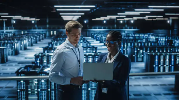 Photo of Data Center Female System Administrator and Male IT Specialist talk, Use Laptop. Information Technology Engineers work on Cyber Security Network Protection in Cloud Computing Server Farm.