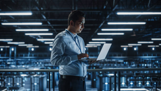 istock Portrait of IT Specialist Uses Laptop in Data Center. Server Farm Cloud Computing Facility with Male Maintenance Administrator Working. Cyber Security and Network Protection. 1336250769