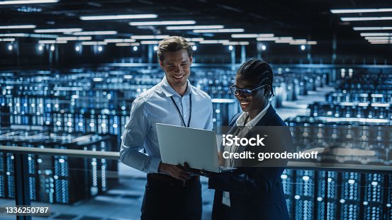istock Data Center Female System Administrator and Male IT Specialist talk, Use Laptop. Information Technology Engineers work on Cyber Security Protection in Cloud Computing Server Farm. 1336250787