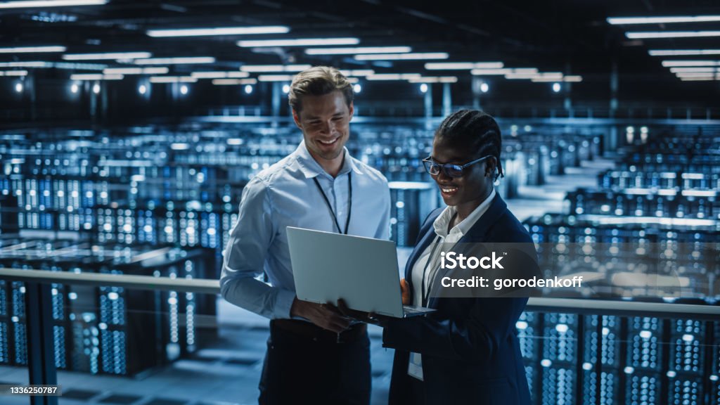 Data Center Female System Administrator and Male IT Specialist talk, Use Laptop. Information Technology Engineers work on Cyber Security Protection in Cloud Computing Server Farm. Technology Stock Photo