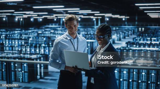 Data Center Female System Administrator And Male It Specialist Talk Use Laptop Information Technology Engineers Work On Cyber Security Protection In Cloud Computing Server Farm Stock Photo - Download Image Now