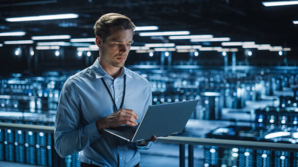 istock Handsome Smiling IT Specialist Using Laptop Computer in Data Center. Succesful Businessman and e-Business Entrepreneur Overlooking Server Farm Cloud Computing Facility. 1336250788