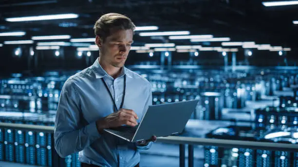 Photo of Handsome Smiling IT Specialist Using Laptop Computer in Data Center. Succesful Businessman and e-Business Entrepreneur Overlooking Server Farm Cloud Computing Facility.